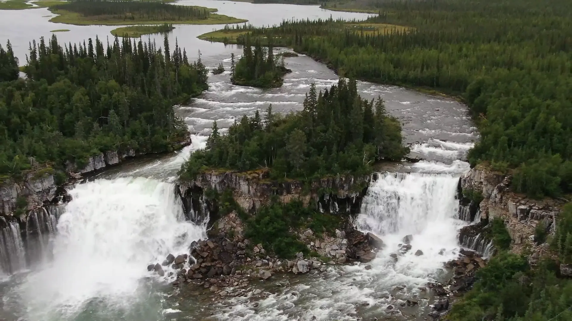 An overhead drone photography image of Whatì Falls. Surrounded by dense forest, the Lac La Martre river narrows upstream, with small rapids visible before being split by a small island to form two 15-metre drops off a rocky cliff. The white water churns against the rocky floor, with small streams of water pouring out of the cliff's walls.