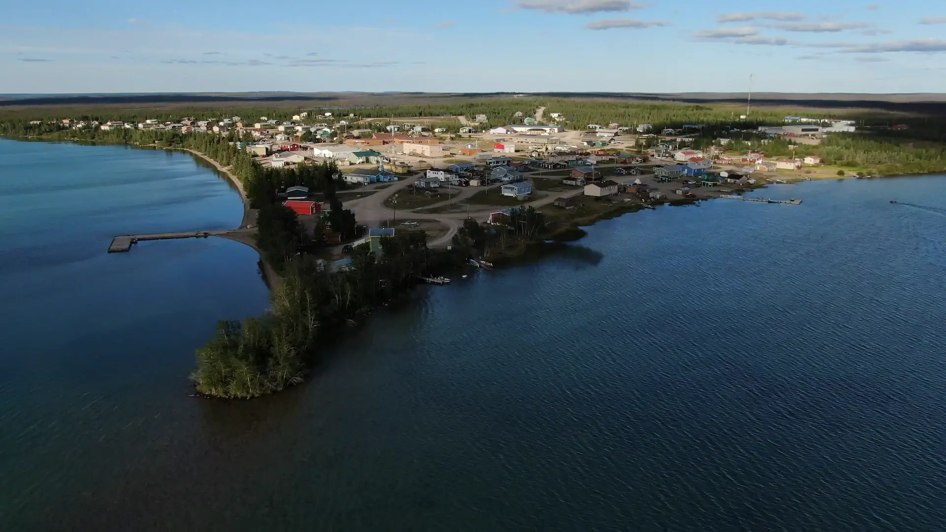 A drone photography image of the clear blue waters of Lac La Martre and the Tłı̨chǫ community of Whatì. Dirt roads connect houses and community buildings in this small town with a population around 500. The colorful buildings dot the town with no particular layout.