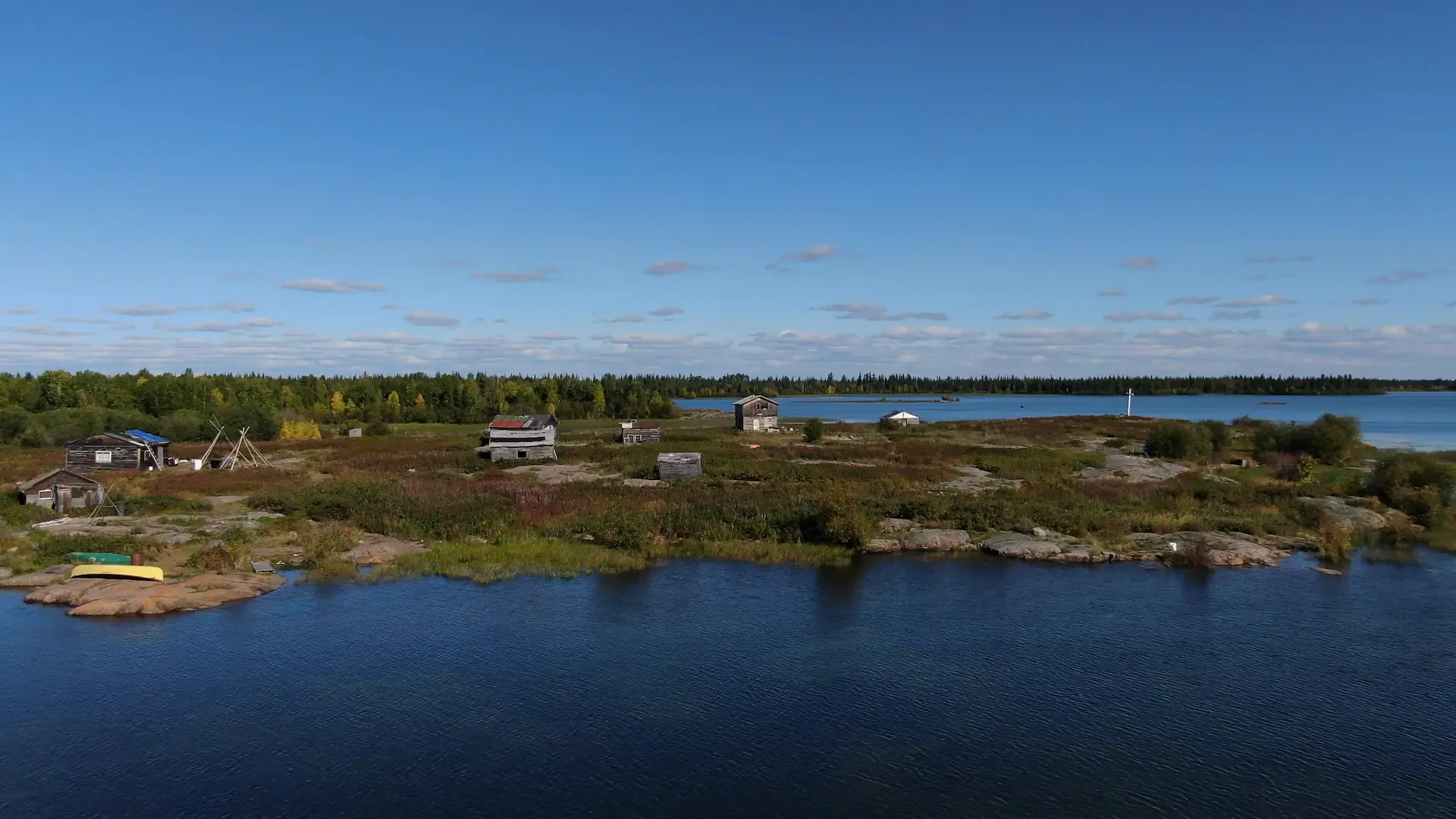 A drone photography image of a small village located on the north shore of Lac La Martre. There are seven small wooden buildings, each made of sun-bleached wood, with patchwork materials used as siding and roofing. The village is located on a patch of rocky land on the edge of Marian Lake.