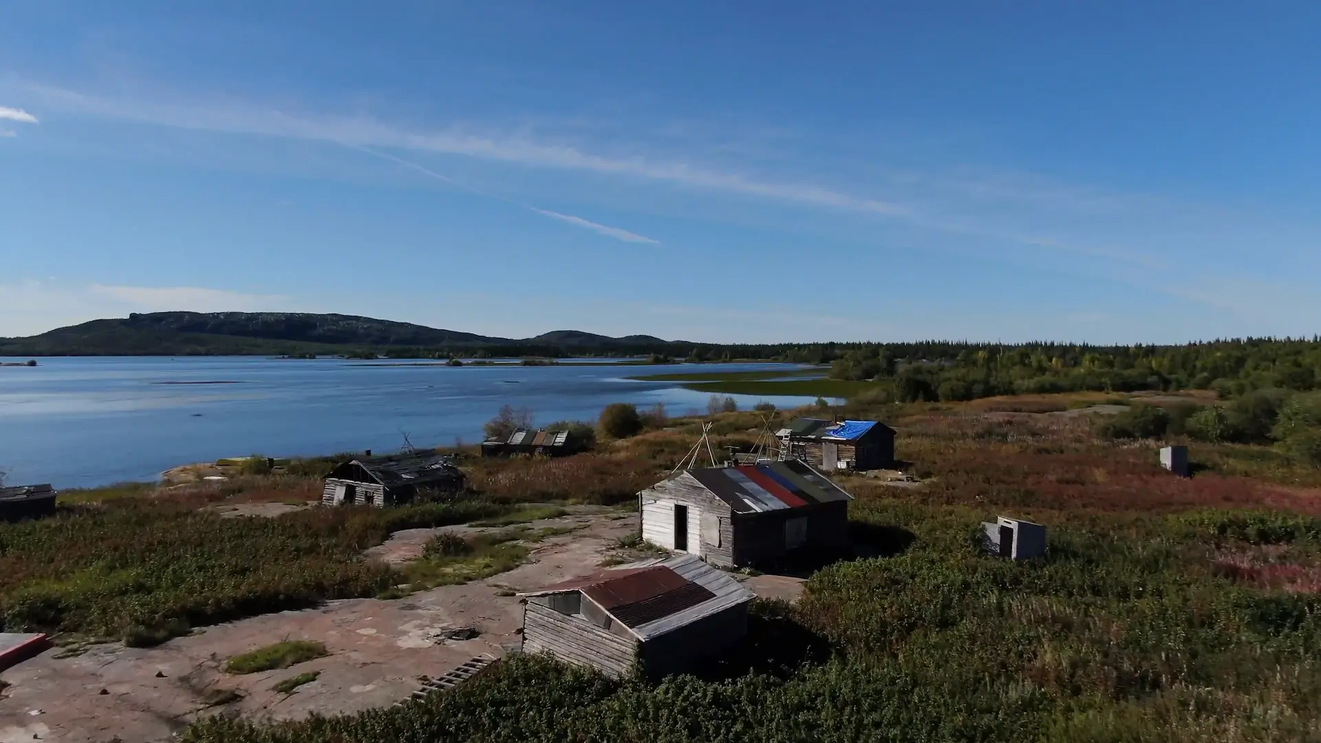 An overhead drone photography shot of a old trading shop located in Marian Village, on the edge of Marian Lake. The roof is made of patchwork materials, and the wood is sunbleached and grey. A low mountain is visible in the distance.