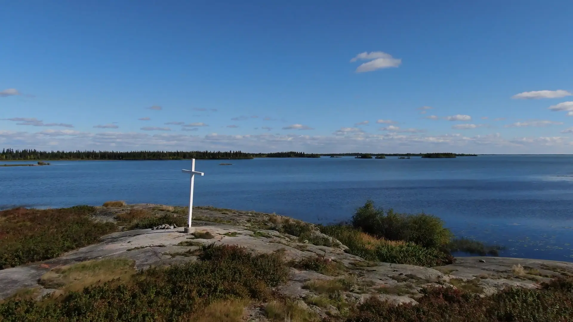 An overhead drone photography shot of the location of a burned down cabin located in Marian Village. A large white cross has been erected on a rock near the shore, overlooking Marian Lake.