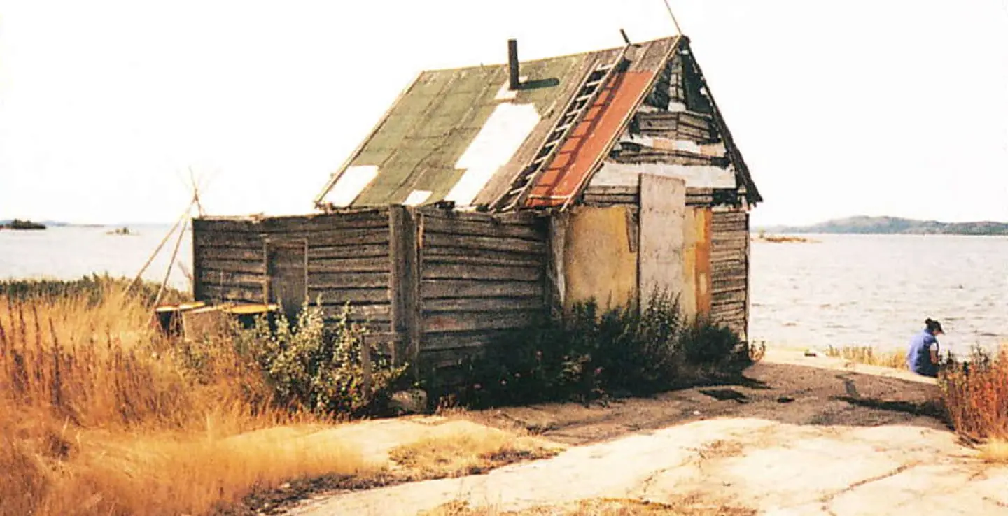 An archival photo of a rundown cabin in Xaelı K'ogola (Marian Village) on the shores of Marian Lake, with boarded up windows, and an old ladder on a patched-up roof. 