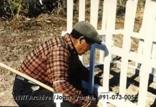 A man in a brown plaid shirt and grey cap kneeling to repair the white picket fence around the grave.