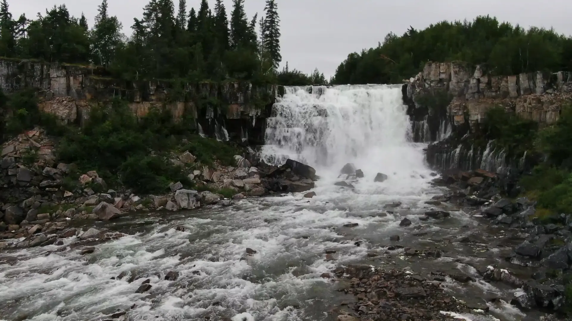 An aerial view of the Lac La Martre river plunging over one of the two 15 foot drops that make up Nàı̨lı̨ı̨ (Whatì Falls). Smaller streams of water pour out of the rocky walls on each side of the river.